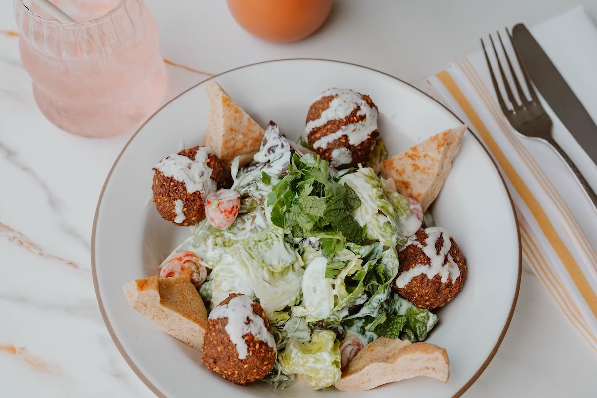 A salad with a creamy dressing, tomatoes, triangles of pita bread and falafel. A light pink drink is next to the plate.