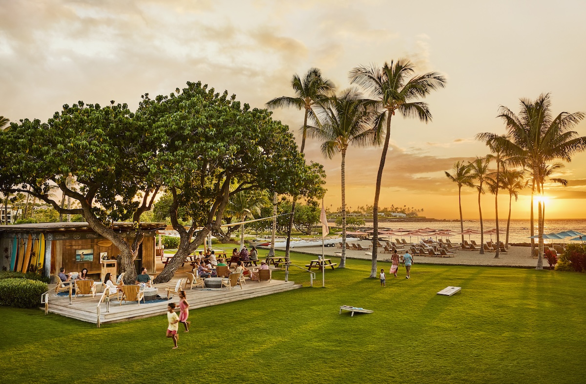A grassy area with outdoor seating, beachfront views, trees, families and a sunset.