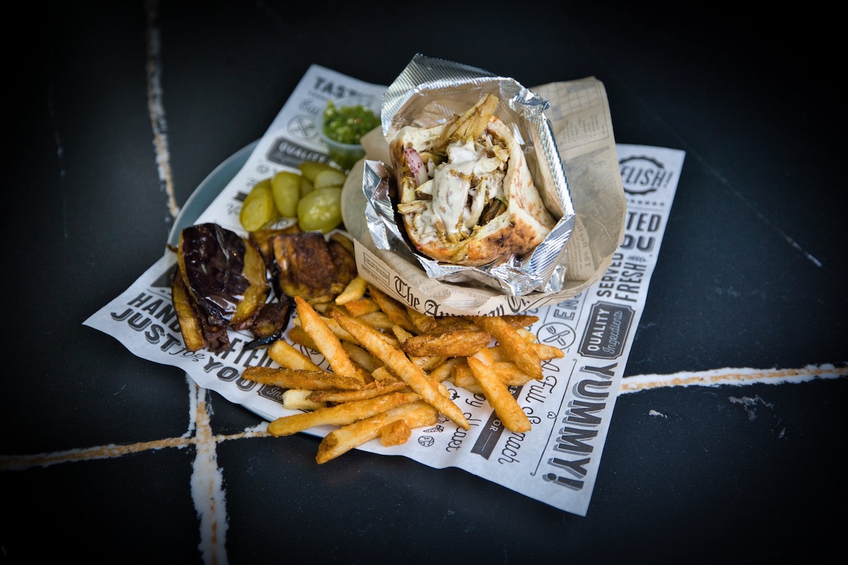 A graphically designed paper with shawarma in a pita and foil and sides including fries and pickles.