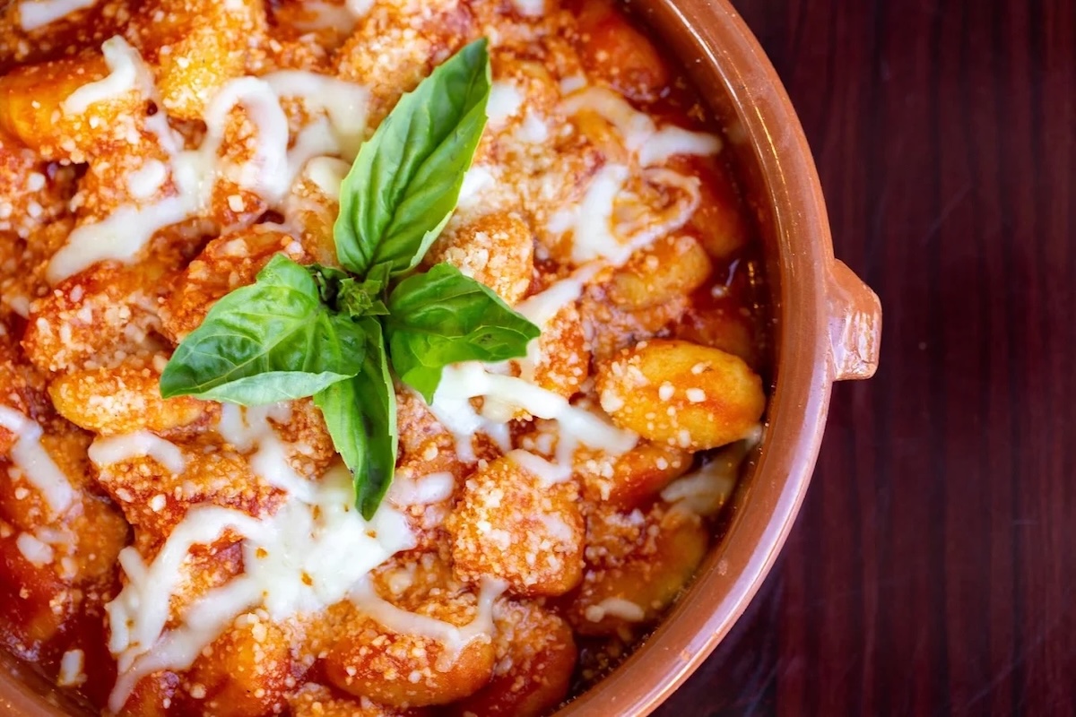 A terracotta colored pot with gnocchi in red sauce and topped with a cheese drizzle and basil for garnish.