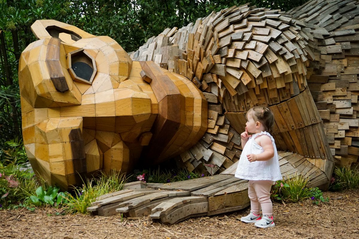 A toddler in a white top stands thoughtfully before the massive wooden face and hand of a reclining troll sculpture nestled in plants.
