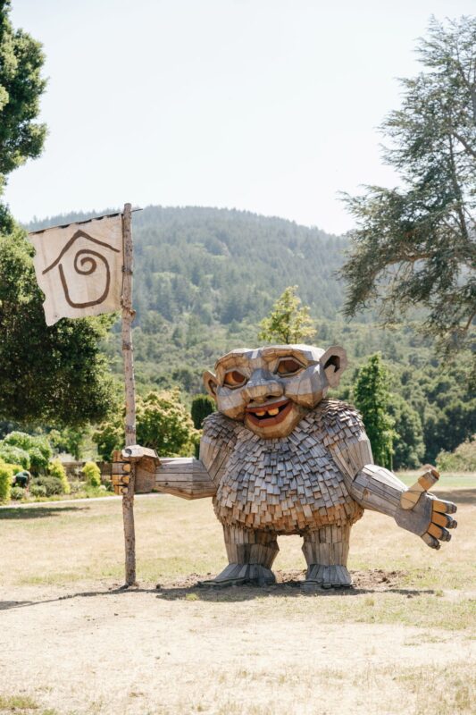 A cheerful troll sculpture made of reclaimed wood holds a flag with a spiral house symbol, standing in a sunlit open field surrounded by trees.