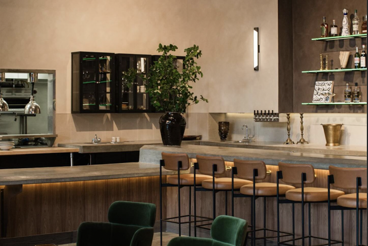 The interior of a restaurant, with bar stools, emerald green chairs and a bar.