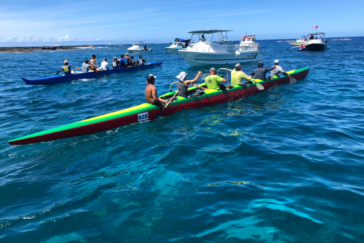 Multiple colorful outrigger canoes with paddlers glide across turquoise waters surrounded by support boats, near a rocky coastline.