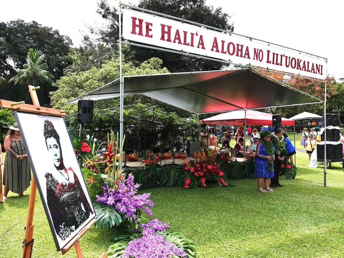 A cultural celebration at Liliuokalani Gardens with a large portrait of Queen Liliuokalani displayed beside a stage decorated with flowers and instruments, under a banner reading “He Hali‘a Aloha No Lili‘uokalani.”