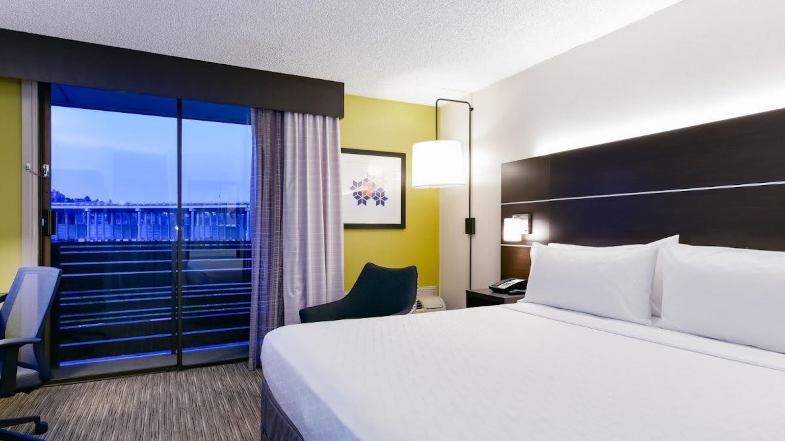 A hotel room with a bed, lamp in the corner, green accent wall and a large window.