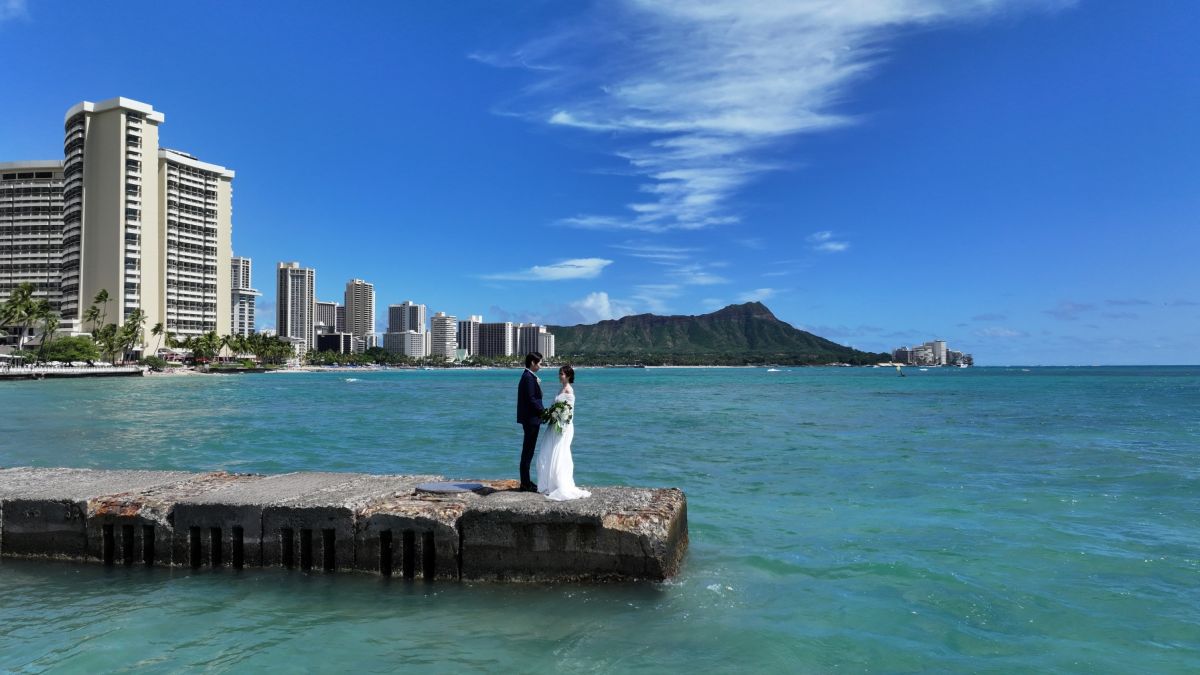 Newlyweds stand on a pier in Waikiki with Diamond Head and city skyline in the background.