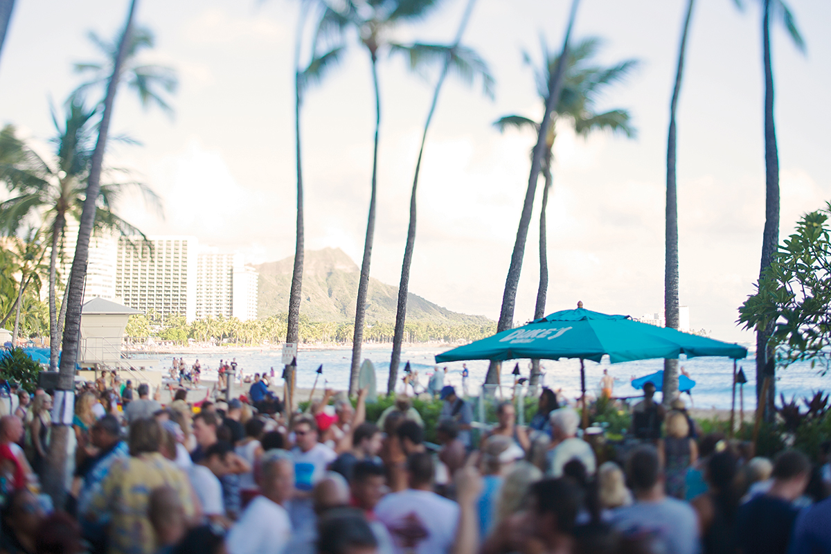 A lively beach scene featuring a crowd of people enjoying the sun, surrounded by tall palm trees.
