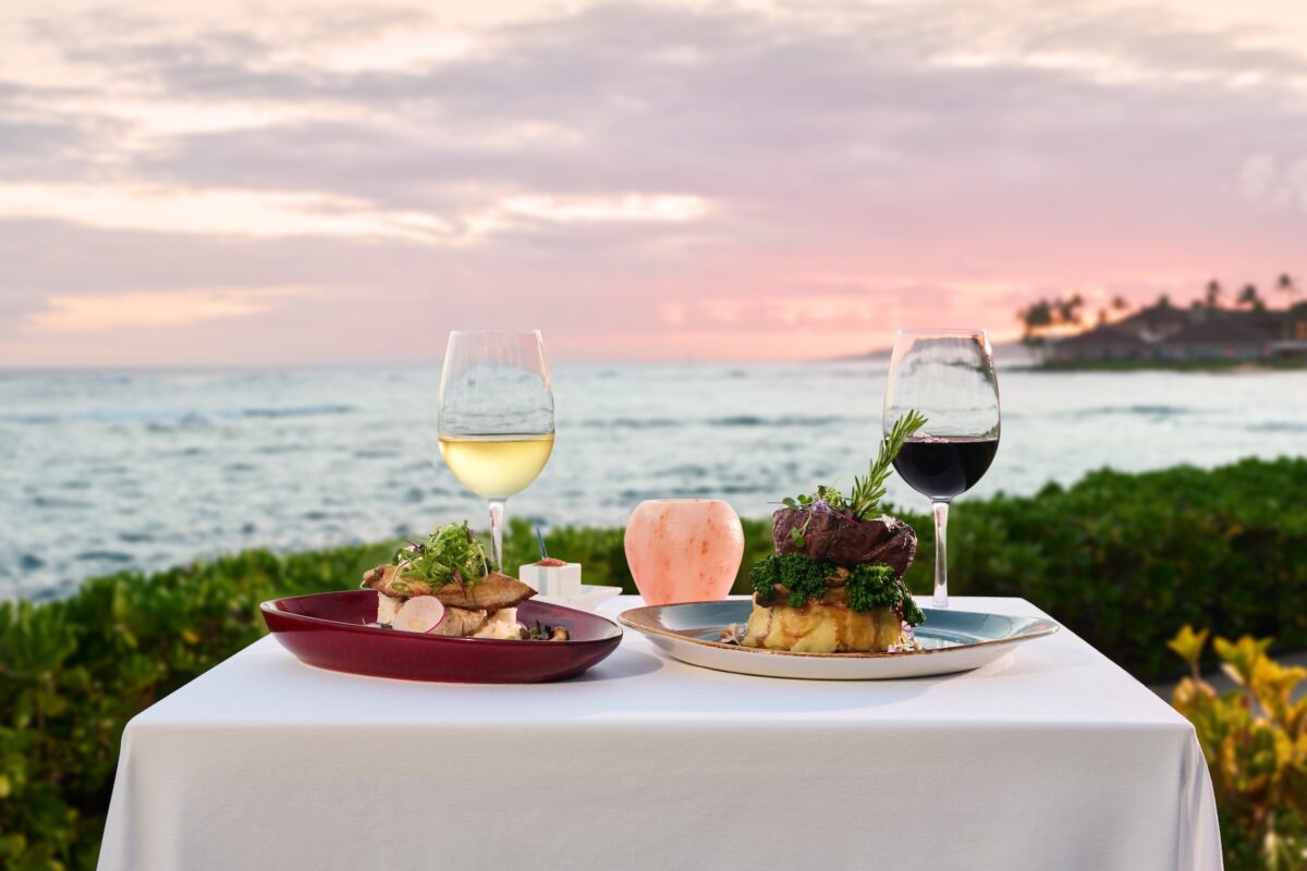 A table with plates of food and glasses of wine overlooking a sunset on the water.