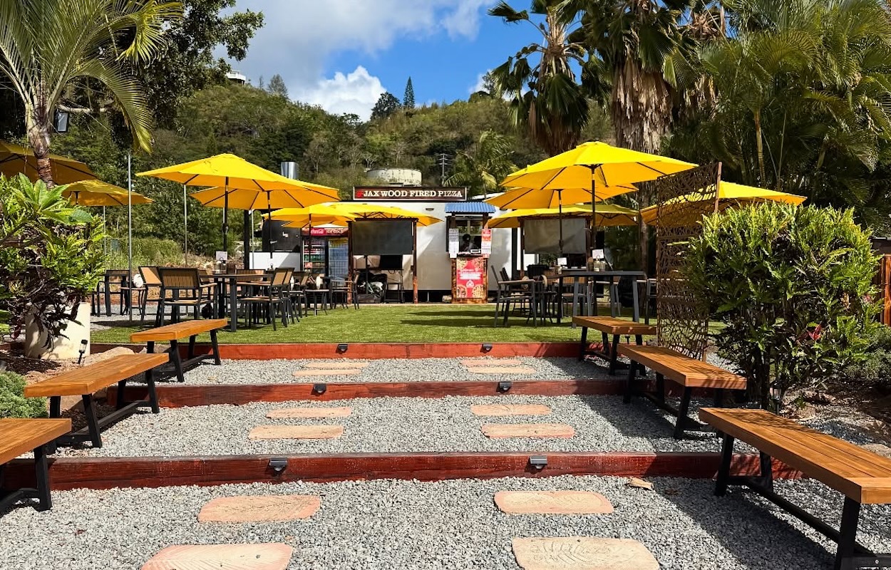 The outdoor setting of a food truck with benches on pea gravel and yellow umbrellas.