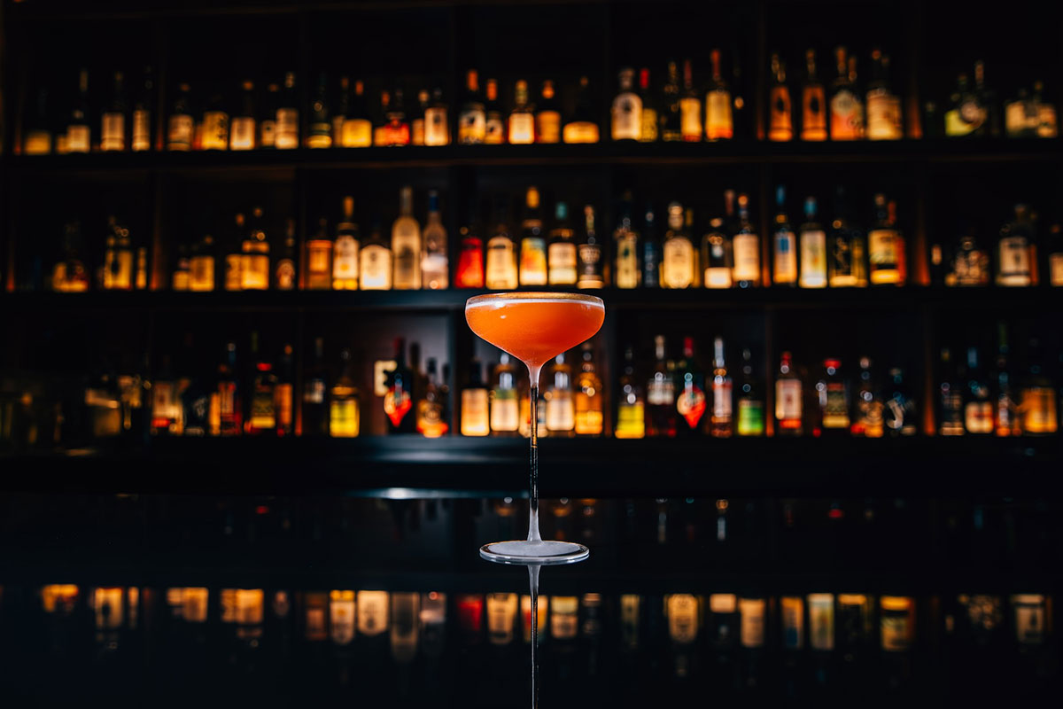 A colorful cocktail placed on a bar counter, surrounded by a wall filled with assorted bottles of spirits.