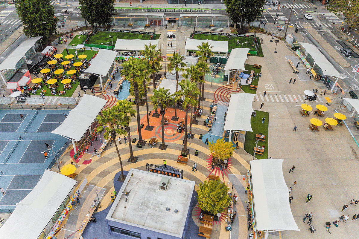 Aerial view of The Crossing at East Cut in San Francisco, featuring palm trees, outdoor seating with yellow umbrellas, sports courts, shaded walkways, and people enjoying the open community space.