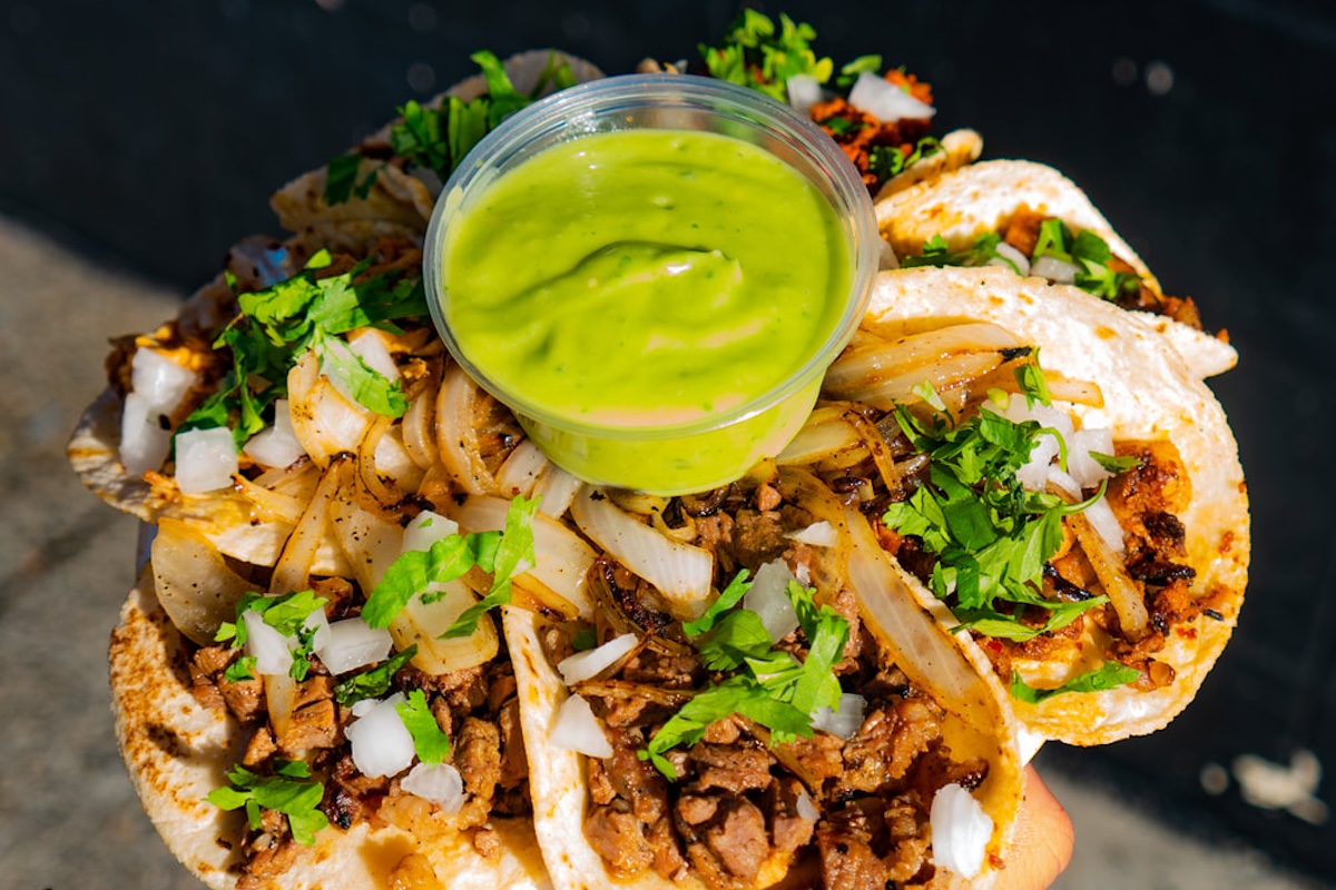 An array of tacos with marinated meat, onions and herbs and a container of bright green sauce in the center.