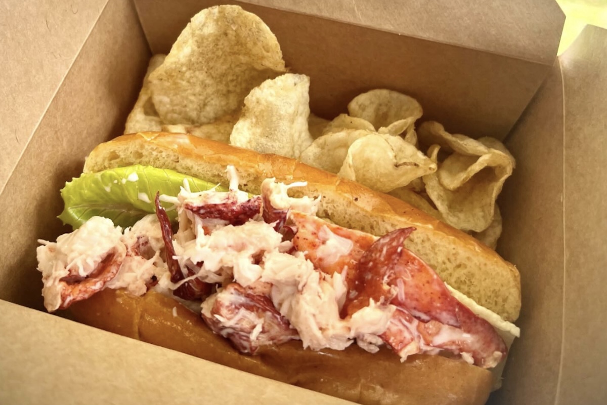 A lobster roll in a paper box and chips on the side.