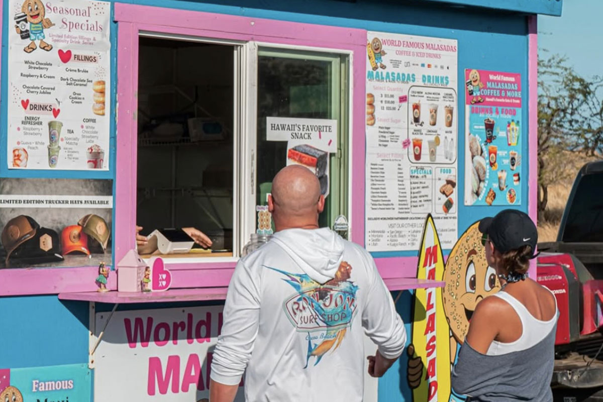 A man and woman stand together in front of a cheerful pink and blue food truck, ready to enjoy some treats.
