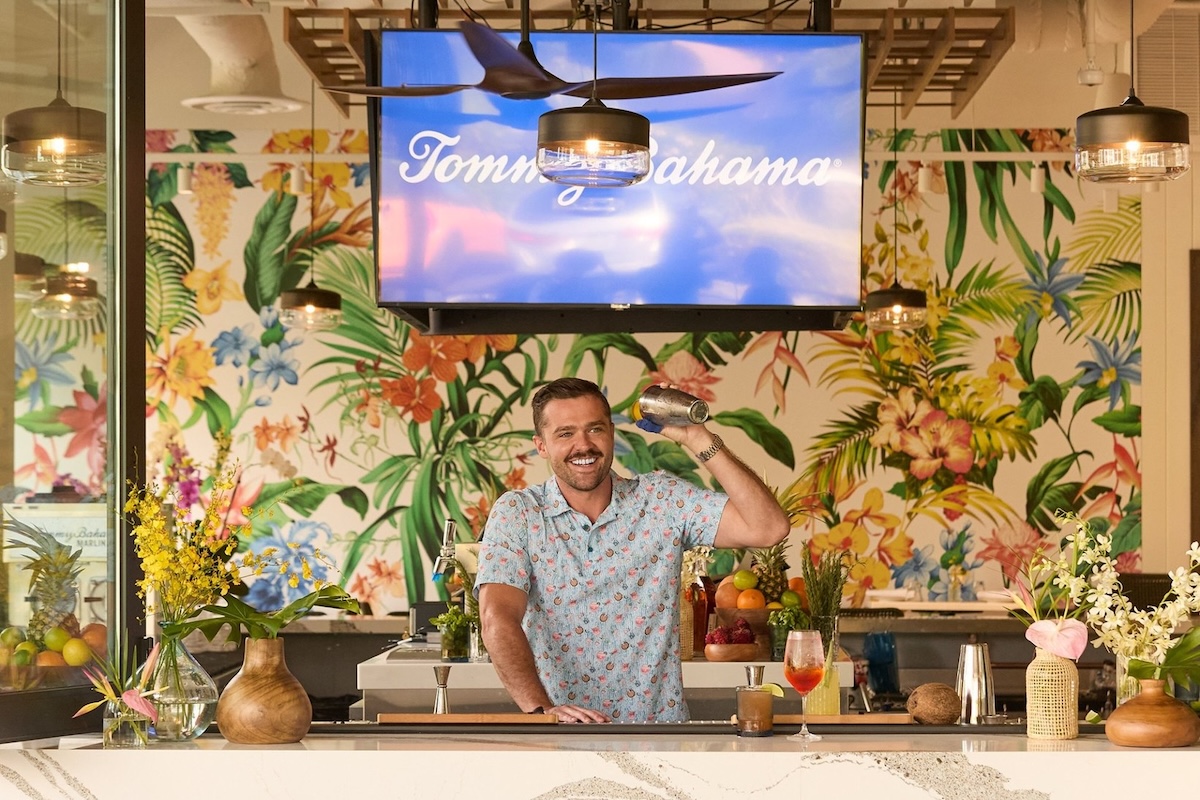 A man wearing a patterned shirt shakes a cocktail shaker with a botanical patterned wallpaper in the background.