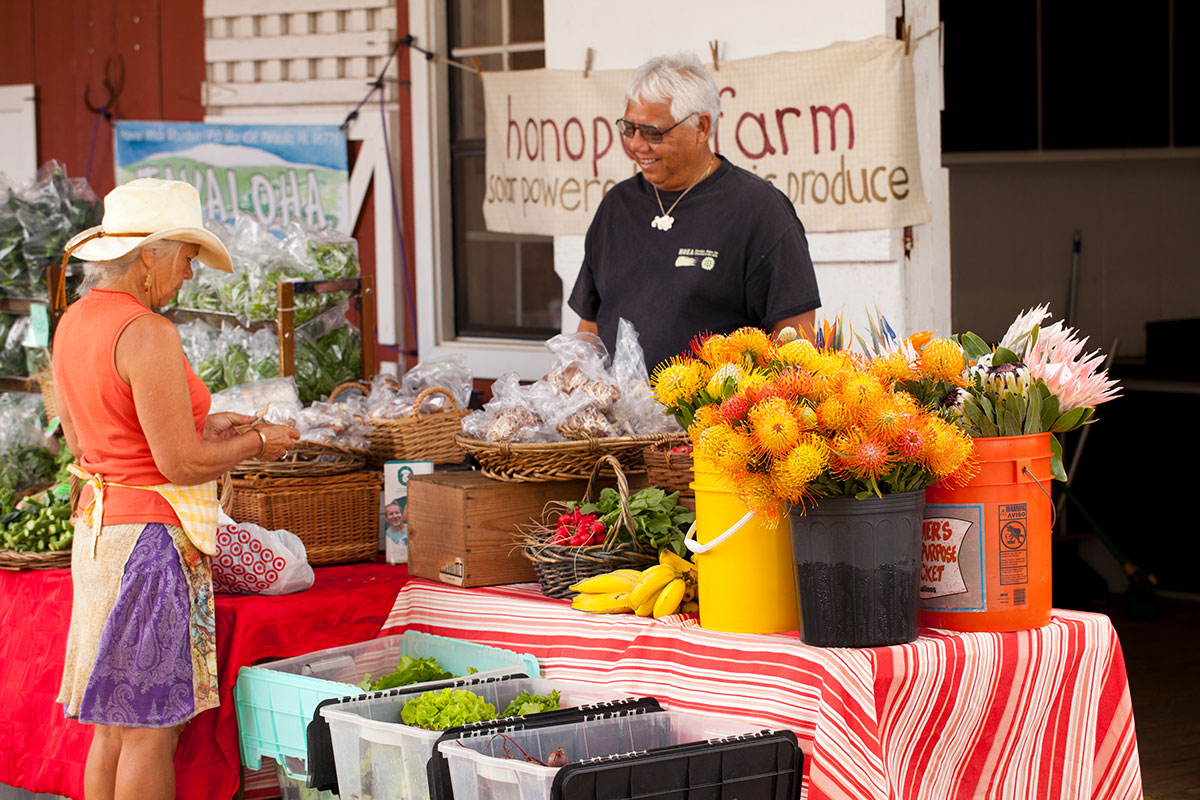A man and woman stand at a table market filled with various fruits and flowers, smiling and engaging with each other.