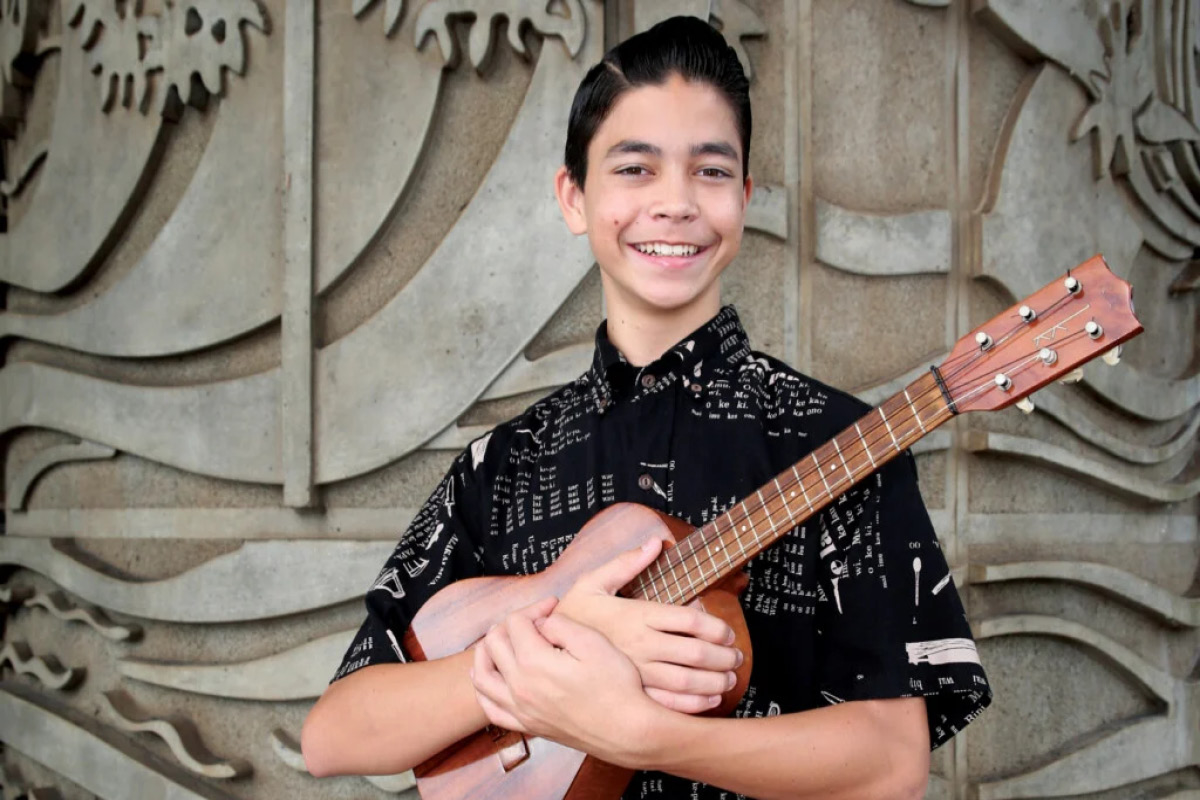 Kamaha‘o Haumea-Thronas stands in front of a wall, holding a ukulele and smiling at the camera.