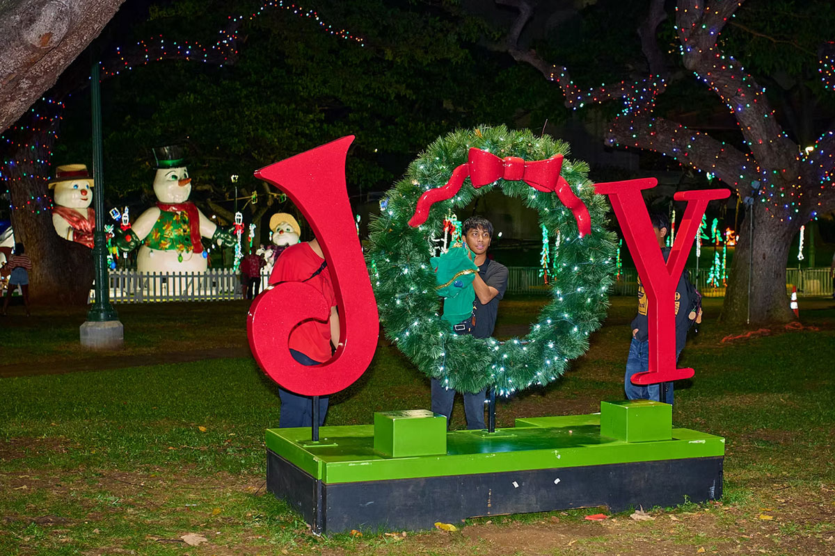 People posing with a large illuminated holiday display spelling ‘JOY,’ featuring a wreath as the ‘O,’ surrounded by Christmas lights and oversized snowman decorations at a festive outdoor event.