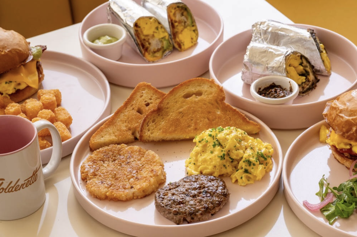 A table spread featuring plates of breakfast food including toast, eggs, sausage, burritos and more.