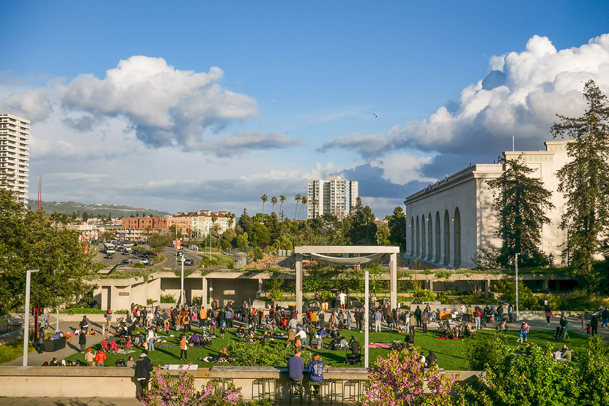 A vibrant outdoor gathering in a sunny urban park, surrounded by lush greenery and flowers.
