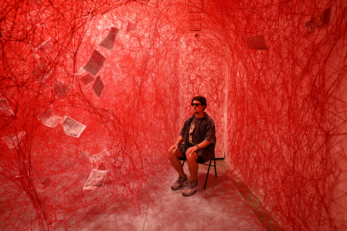 A person sits in a chair in a red art installation filled with tangled threads and scattered papers, creating an intense, immersive atmosphere.
