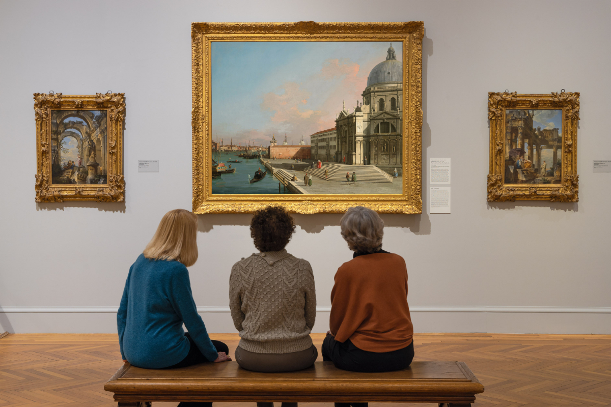 Three people sit on a bench in an art gallery, viewing a large painting of a Venetian scene.