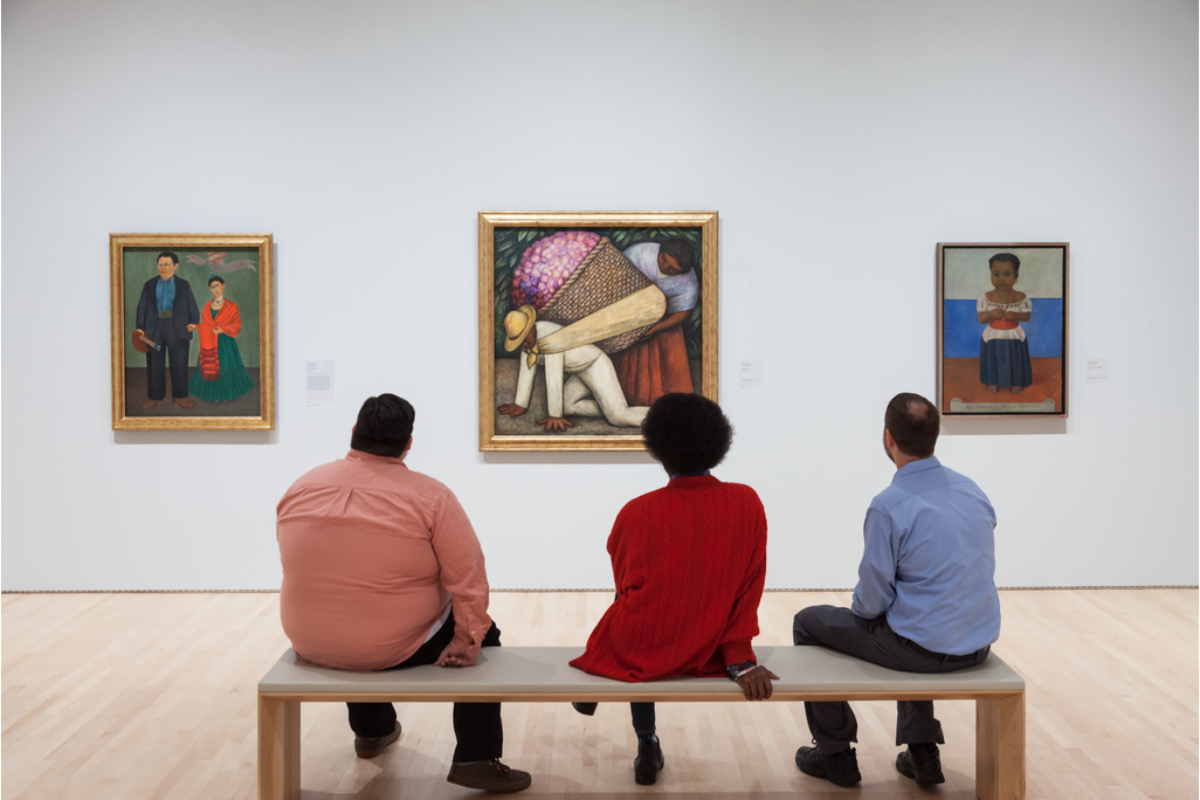 Four people sit on a bench in a gallery, viewing three framed paintings on a white wall.