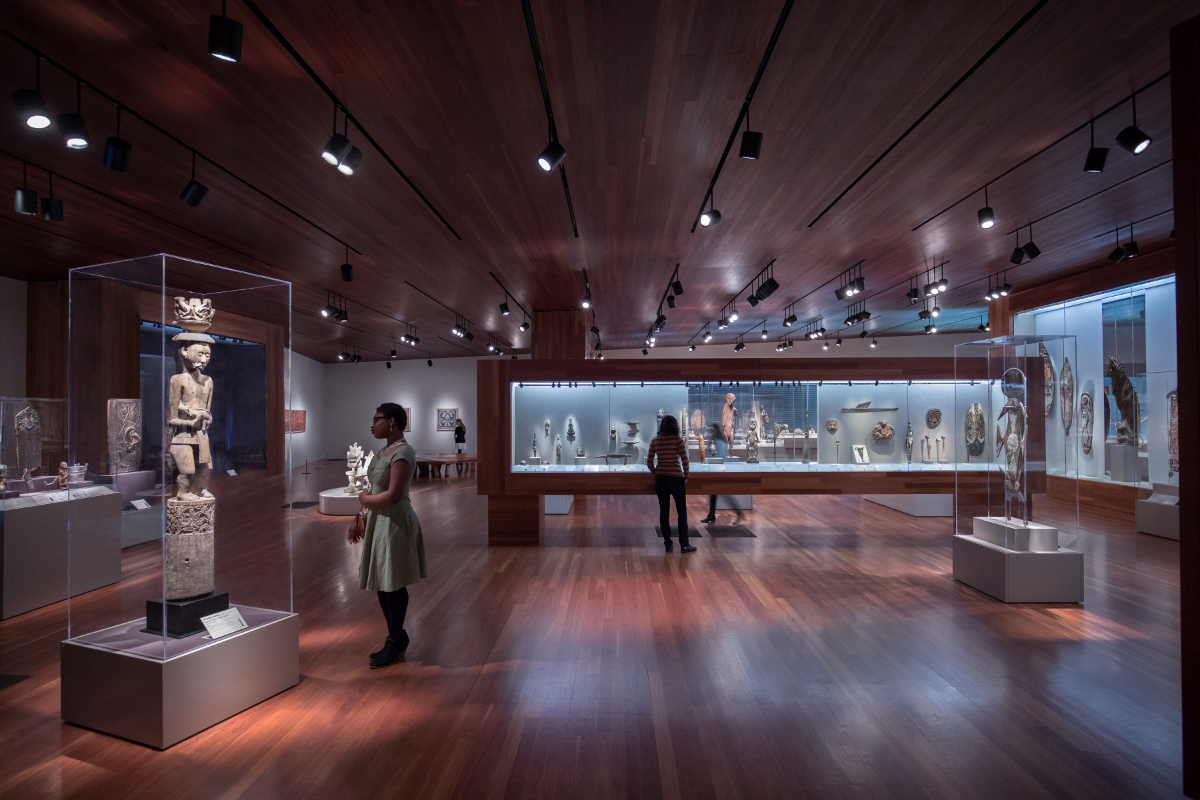 A spacious art gallery with wooden floors and high ceilings, displaying cultural sculptures in glass cases.