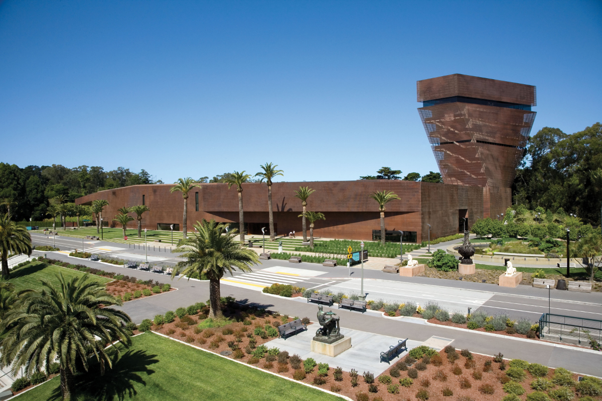 A modern, angular museum with a rust-colored exterior is surrounded by lush greenery, palm trees, and landscaped gardens under a clear blue sky.