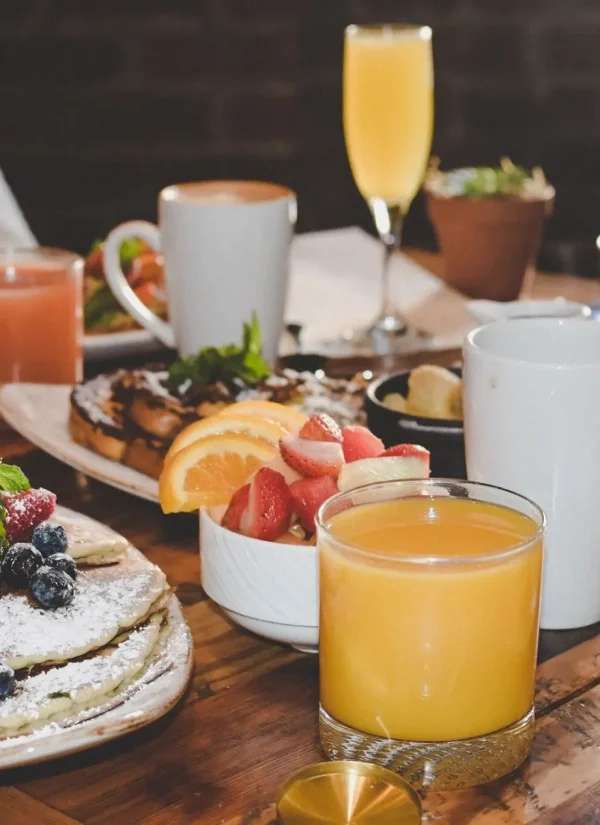 A table set for brunch with pancakes topped with berries and powdered sugar, a bowl of sliced fruit, various drinks including orange juice and coffee, and an assortment of other breakfast dishes. A hand pours syrup onto the pancakes.