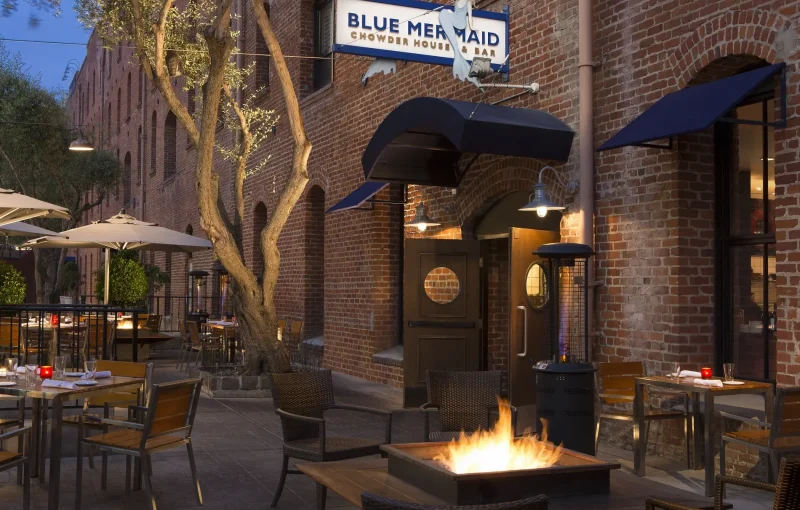 Outdoor dining area of a restaurant called "Blue Mermaid Chowder House & Bar" with brick walls, an arched entrance, tables and chairs, and a central fire pit. The setting appears cozy with ambient lighting and umbrellas over some tables.