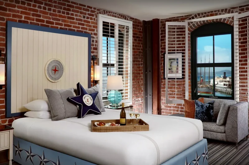 A cozy bedroom featuring exposed brick walls, a large bed with nautical-themed decor, and a cushioned armchair by the window. A tray holding wine and glasses rests on the bed. The window offers a view of a waterfront with boats and a clear sky.