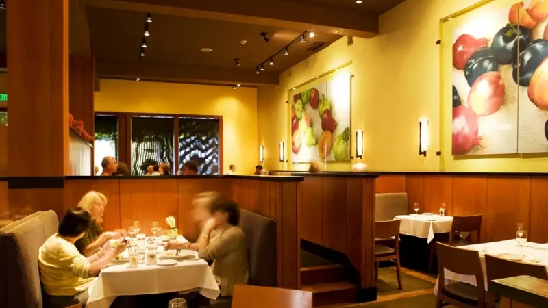 A cozy restaurant interior with soft lighting and wooden furnishings. Diners are seated at tables with white tablecloths, enjoying their meals. Colorful fruit paintings adorn the warm yellow walls. There's a partially visible window with outdoor views.