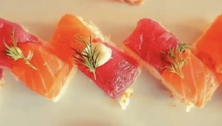 Close-up of rectangular pieces of sushi topped with slices of raw salmon and tuna, garnished with small sprigs of dill and a touch of wasabi or cream. The sushi pieces are arranged in a zigzag pattern on a light-colored plate.