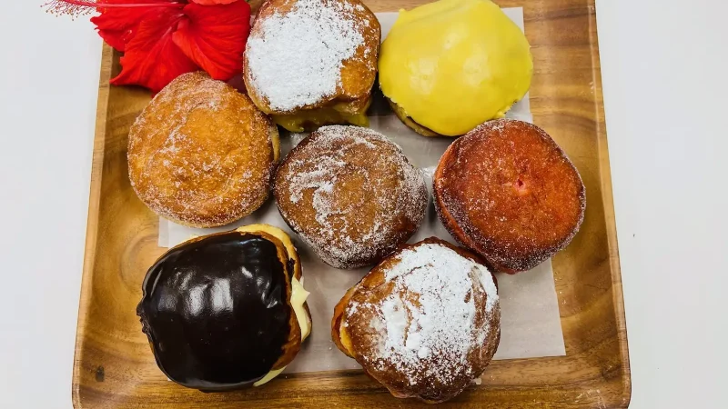 An assortment of seven colorful doughnuts arranged on a wooden square plate, each topped with different glazes and powdered sugar. A small bunch of vibrant red hibiscus flowers is placed in the upper left corner of the plate for decoration.