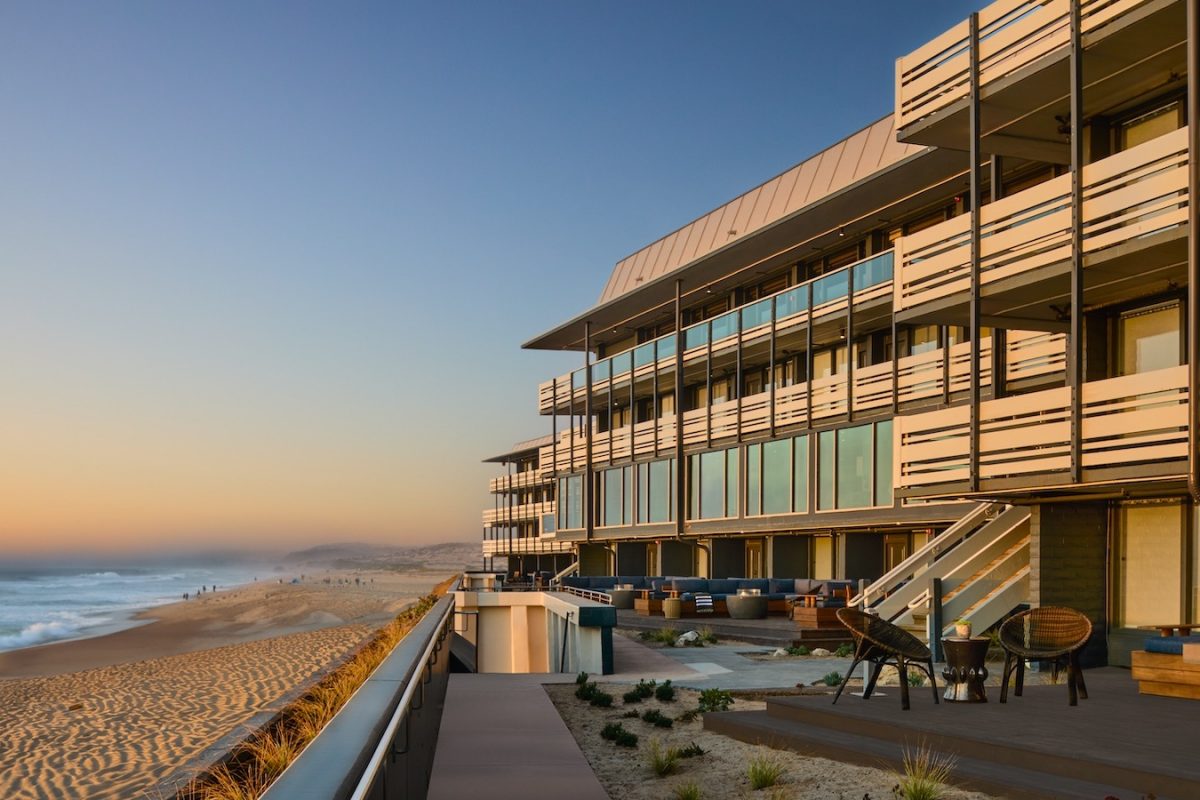 Modern beachfront building with balconies and large windows overlooking a sandy beach at sunset. The boardwalk of Monterey Beach Hotel features outdoor seating areas and planters, and the ocean's waves gently lap against the shore. Distant figures are seen walking along the beach.