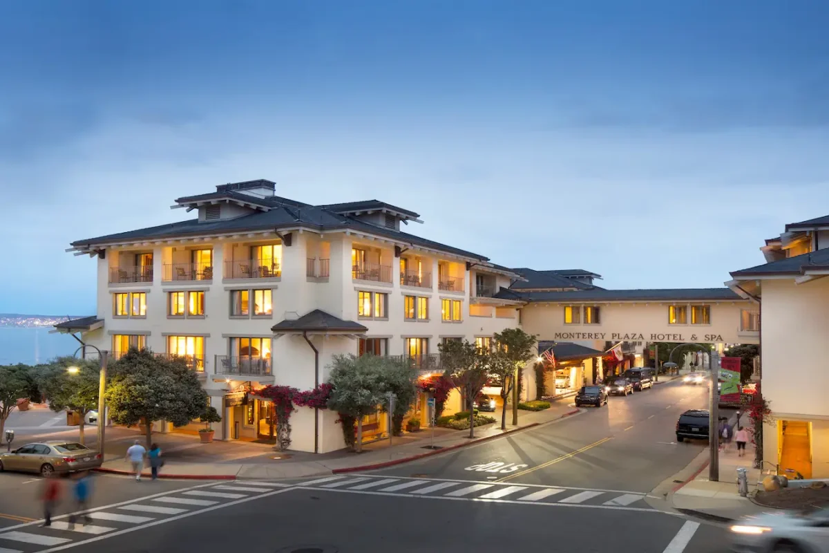 Street view of the Monterey Plaza Hotel and Spa at dusk. The well-lit building features illuminated windows, with people strolling along the sidewalks. Nearby, plants and flowers adorn the exterior, while the ocean shimmers in the background.