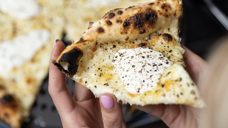 Pizzana-Los Angeles-@pizzana Hand holding a slice of charred, wood-fired pizza with melted cheese, black pepper, and a dollop of ricotta or creamy topping.