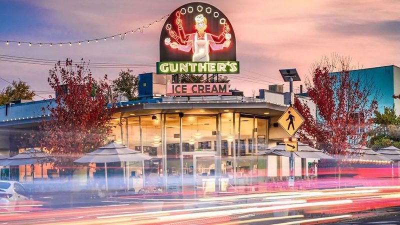 A brightly lit ice cream shop named Gunther's, featuring a neon sign with a smiling ice cream figure. Known for offering the best dessert in Sacramento, the building is surrounded by trees with autumn foliage, outdoor seating, and string lights during sunset. Blurred car lights streak across the foreground.