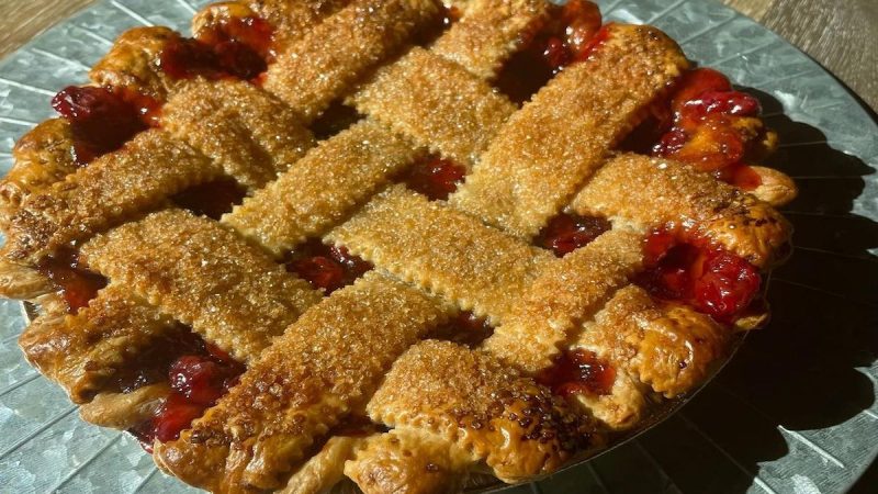 A golden-brown lattice-topped pie filled with vibrant red cherry filling sits on a decorative metal plate. The crust, sprinkled with sugar, glistens under natural light, highlighting its homemade appeal. Known as the best dessert in Sacramento, it’s a treat you can't miss!