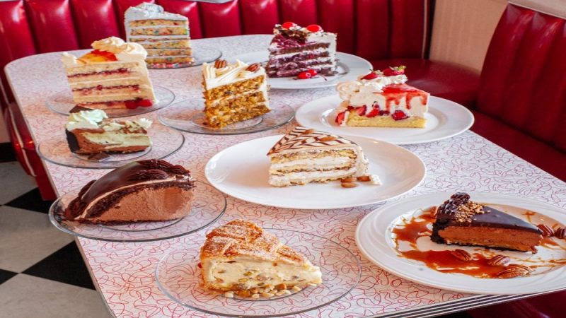 A variety of plated cakes and slices, considered the Best Dessert in Sacramento, are arranged on a retro-style pink and white table with red vinyl booth seating. The cakes range in type and decoration, including chocolate, layered, cream-topped, fruit garnished, and drizzled with sauces.