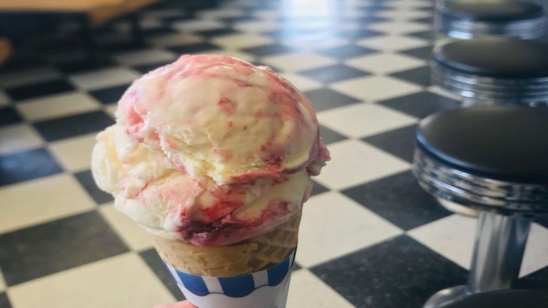 A hand holds a paper-wrapped ice cream cone topped with a scoop of vanilla ice cream swirled with raspberry. The Best Dessert in Sacramento is savored at an ice cream parlor in the background, featuring black and white checkered flooring, chrome barstools with black seats, and wooden booths.