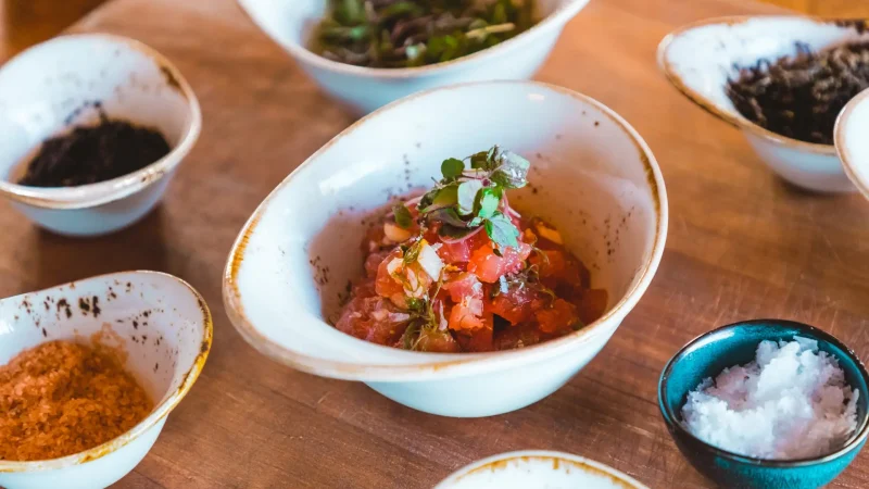 A close-up of various dishes in white bowls on a wooden surface. The central bowl contains a colorful mixture of diced vegetables and greens. Surrounding bowls hold different condiments and ingredients, including coarse salt and herbs. The setting appears casual.