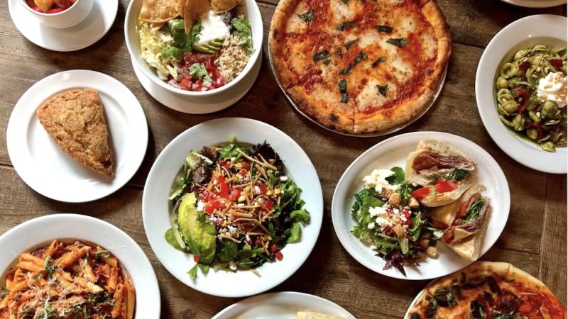 Zinc Cafe and Bar-Los Angeles-@zinccafeandbar Top-down view of a rustic table covered in various dishes including pizzas, pasta, salads, a burrito, a slice of pie, and a cup of fruit.