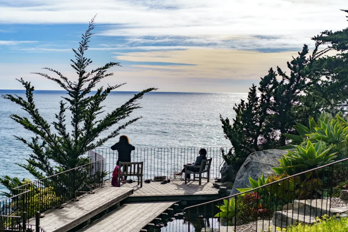 Two people enjoy a scenic ocean view from an elevated wooden platform. One stands by the railing while the other sits on a chair, surrounded by lush greenery and trees. The sky is partly cloudy, and the ocean is calm, mirroring the tranquil ambiance found at many Big Sur hotels.