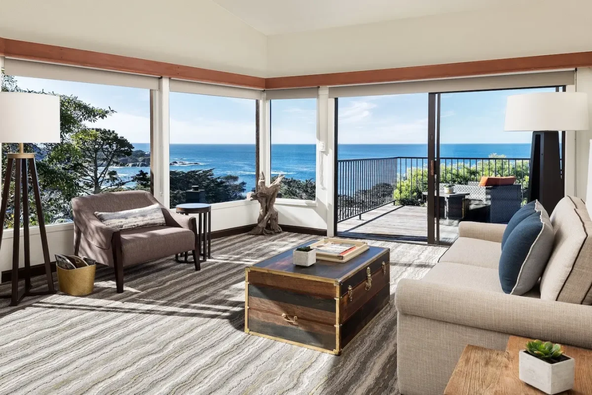 A cozy, sunlit living room with large windows and a sliding glass door leading to a balcony with an ocean view. The room, reminiscent of the luxurious accommodations found at Carmel Group Hotels, features a beige sofa, a wooden coffee table, an armchair, and modern lamps. A coastal landscape and blue sky are visible outside.