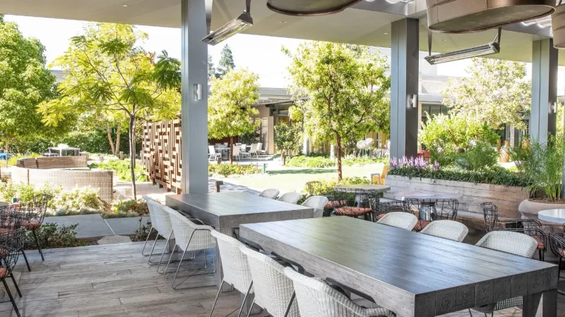 A spacious outdoor restaurant with wooden tables and white, casual seating under a shaded patio. Surrounding the area are lush green trees, potted plants, and garden beds. The setting is serene with a mix of sun and shade in a modern, garden-like environment.