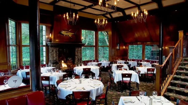 A warmly lit, elegant restaurant with round tables covered in white tablecloths, each set with wine glasses and silverware. The room features large windows, wooden walls, a cozy stone fireplace, and chandeliers. A staircase is visible on the right.