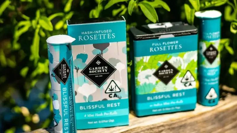 Assorted Garden Society cannabis products, including hash-infused pre-rolls and full-flower rosettes, are displayed outdoors on a wooden surface surrounded by lush green foliage. The packaging, reminiscent of travel sleep essentials, features a blue, white, and green color scheme.